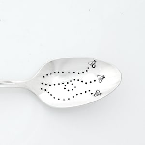 May include: A silver spoon with a floral design on the handle and the words "si sapis sis apis" engraved on the handle. The bowl of the spoon has a pattern of dots and three bees.