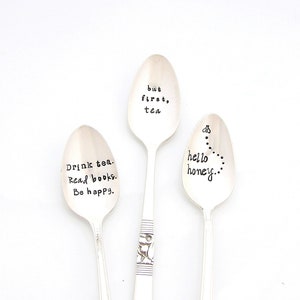 May include: Three silver spoons with engraved sayings. The spoons have a floral design at the end of the handle. The sayings are "but first, tea", "Drink tea. Read books. Be happy." and "hello honey..."