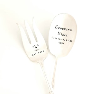May include: A set of two silver-plated serving utensils, a fork and a spoon. The fork has the initials "ELB" and the date "Est. 2023" engraved on it. The spoon has the text "Spooning Since October 7, 2023" engraved on it.