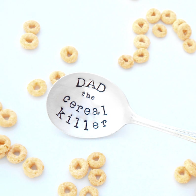 May include: A silver spoon with the text "DAD the cereal killer" engraved on it. The spoon is surrounded by small, yellow cereal rings.