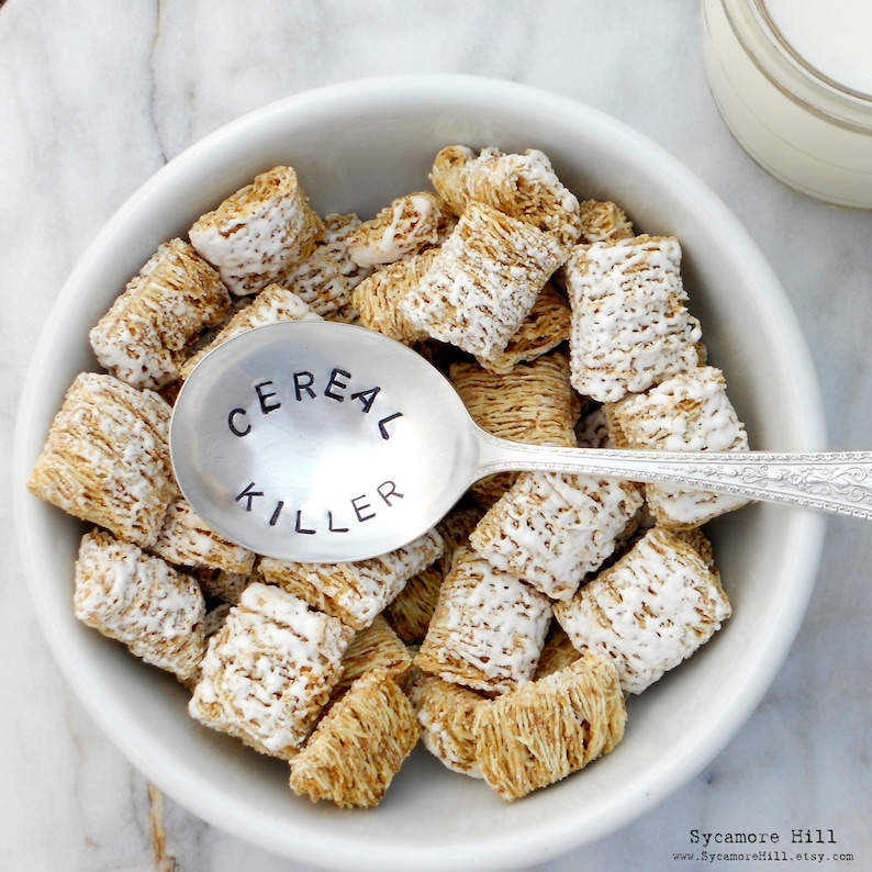 May include: A silver spoon with the words "Cereal Killer" engraved on it, resting in a bowl of frosted cereal.