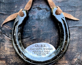 May include: A black horseshoe with a brown leather strap. A silver metal tag is attached to the horseshoe with the text "DUKE 2019 HORSE SPONSOR JACKSON HOLE THERAPEUTIC RIDING".