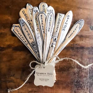 May include: A set of vintage silverware garden markers with names of herbs and vegetables written on them, tied together with twine and a tag that reads "Sycamore Hill The ORIGINAL Louisville Since 2010."