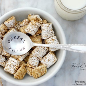 May include: A white bowl filled with frosted mini wheat cereal and a silver spoon with the words "Cereal Killer" engraved on it. The spoon is resting on top of the cereal. The text "The Original Cereal Killer Spoon" is printed below the bowl.
