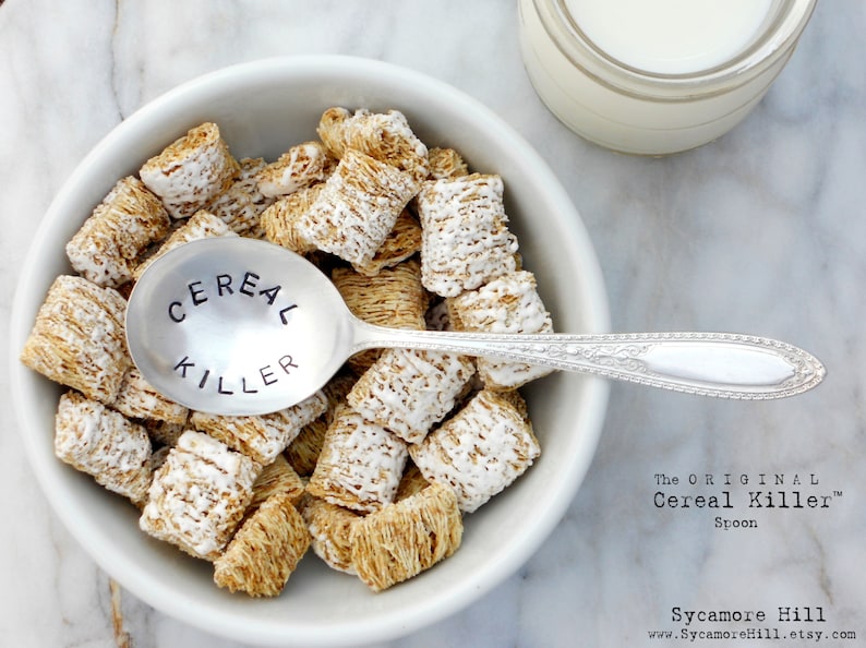 May include: A white bowl filled with frosted mini wheat cereal and a silver spoon with the words "Cereal Killer" engraved on it. The spoon is resting on top of the cereal. The text "The Original Cereal Killer Spoon" is printed below the bowl.