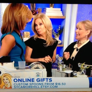 May include: Three women are standing in front of a blue background. The woman on the left is wearing a blue top. The woman in the middle is wearing a black top. The woman on the right is wearing a black jacket with a white collar. The text "ONLINE GIFTS CUSTOM SPOONS FROM &pound;16.50 SYCAMOREHILL.ETSY.COM" is displayed on a white banner.