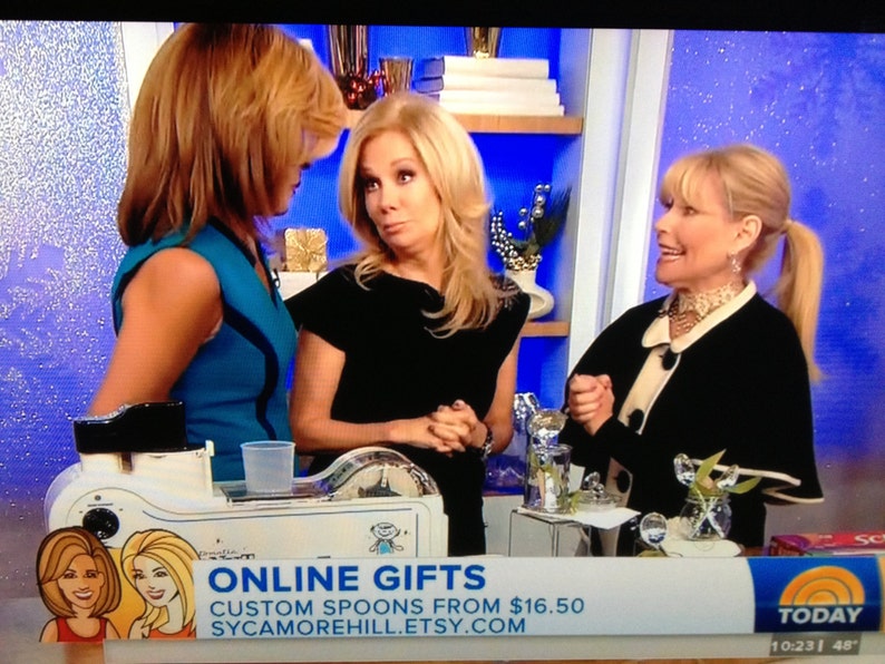 May include: Three women are standing in front of a blue background. The woman on the left is wearing a blue top. The woman in the middle is wearing a black top. The woman on the right is wearing a black jacket with a white collar. The text "ONLINE GIFTS CUSTOM SPOONS FROM &pound;16.50 SYCAMOREHILL.ETSY.COM" is displayed on a white banner.