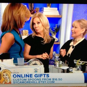 May include: Three women are standing in front of a blue and white background. The woman on the left is wearing a blue and white dress. The woman in the middle is wearing a black dress. The woman on the right is wearing a black jacket with a white collar. The woman in the middle is holding a spoon. The text "ONLINE GIFTS CUSTOM SPOONS FROM $16.50 SYCAMOREHILL.ETSY.COM" is displayed on the bottom of the image.