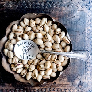 May include: A silver spoon with the text "Keep Your Hands Off My Nuts!" engraved on it. The spoon is lying on top of a bowl of pistachios.