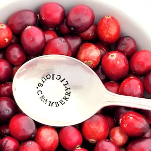 May include: A silver spoon with the words "Cranberrylicious" engraved around the bowl, surrounded by red cranberries.
