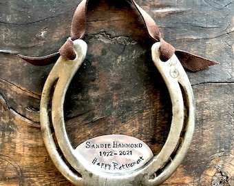 May include: A silver horseshoe with a brown leather strap. A silver plaque is attached to the horseshoe with the text "Sandie Hammond 1972-2021 Happy Retirement".