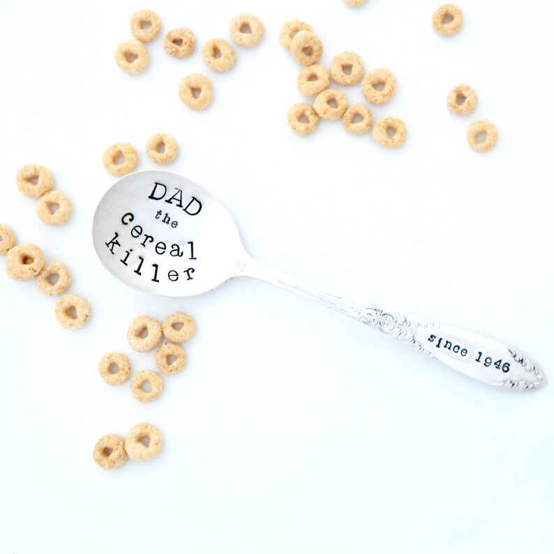 May include: A silver spoon engraved with the text "DAD the cereal killer since 1946". The spoon is surrounded by scattered cereal rings on a white background.
