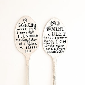 May include: Two silver spoons with black text on them. The text on the left spoon reads "THE Oaks Lily sweet $ sour ICE VODKA cranberry juice and a splash of TRIPLE SEC". The text on the right spoon reads "THE MINT JULEP fresh CRUSHED mint ice LEAVES simple syrup KENTUCKY BOURBON".