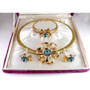 May include: A gold tone jewelry set with a flexible necklace, a brooch, and earrings. The jewelry features blue stones and a bow design.