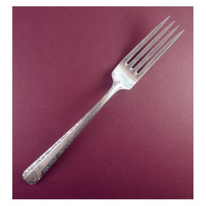 May include: A silver fork with four tines and a long handle. The handle is decorated with an ornate, floral pattern. The fork is set against a burgundy background.