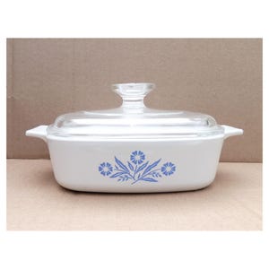May include: White rectangular casserole dish with a clear glass lid. The dish features a blue floral design on the front. The lid has a clear glass handle. The dish is on a neutral surface.