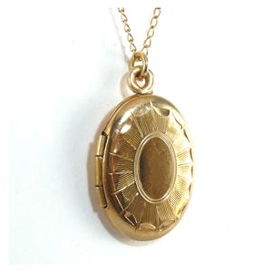 May include: A gold-colored oval locket pendant with a sunburst design, suspended from a delicate gold chain. The locket has a hinged opening and a smooth, oval center. The necklace is displayed against a white background.