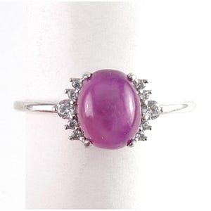 May include: A silver ring featuring a large, oval, purplish-pink gemstone, surrounded by smaller, clear gemstones. The ring's band is thin and polished, with the central stone prominently displayed.