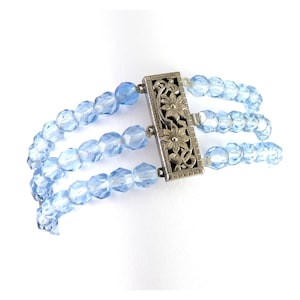 May include: A vintage bracelet featuring three strands of light blue faceted beads. The bracelet is secured by a silver-toned clasp with a floral design. The beads are evenly spaced, creating a delicate and elegant accessory.