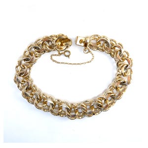 May include: A gold-tone metal chain bracelet featuring a series of interlocking circular links with a textured finish. The bracelet includes a secure box clasp closure and a thin safety chain attached to the side to ensure it stays fastened.
