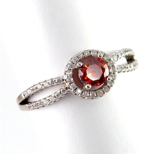 May include: A silver ring featuring a round, fiery red gemstone encircled by a halo of smaller, clear stones. The band splits into two, each lined with additional clear stones, creating a sparkling, elegant design.