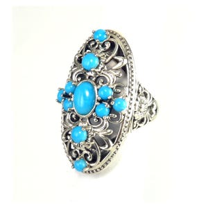 May include: A silver-toned ring with an oval turquoise stone at the center, surrounded by smaller turquoise stones and ornate filigree detailing. The ring has a floral design on the band and a bright, polished finish.