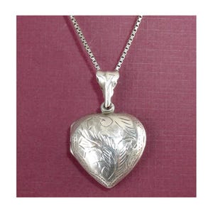 May include: A silver heart-shaped locket pendant with intricate floral engraving, suspended from a delicate chain. The locket is closed, and the design is symmetrical. The background is a solid maroon color.