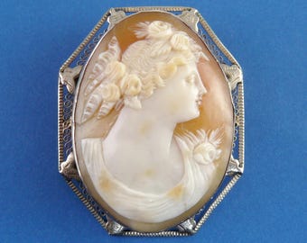 May include: Oval cameo brooch featuring a carved profile of a woman with floral accents. The cameo is set in a silver-toned filigree frame. The woman's profile is in a cream color, set against a light orange background.