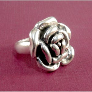May include: A silver ring featuring a detailed rose design. The ring's floral element is fully sculpted, with petals arranged to mimic a blooming rose. The ring is set against a burgundy background, highlighting the silver's shine.