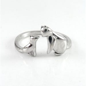 May include: A silver-colored ring featuring a cat and a penguin design. The ring has a split shank design, with the cat and penguin facing each other. The ring is made of a shiny, metallic material.