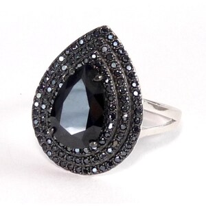 May include: A ring featuring a large, pear-shaped black center stone surrounded by two concentric halos of small, round black stones. The setting is dark metal, and the band is a simple, polished silver-toned metal that splits into two parts.