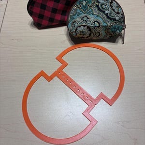May include: Two small zippered pouches and an orange plastic template. One pouch features a red and black buffalo plaid pattern, while the other has a teal, black, and cream paisley print. The orange template is a flat, symmetrical shape.
