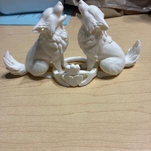 Può includere: Una scultura bianca stampata in 3D raffigurante due lupi che ululano. I lupi sono posizionati su entrambi i lati di una base a forma di cuore con una corona. La scultura è realizzata in un materiale di colore chiaro.