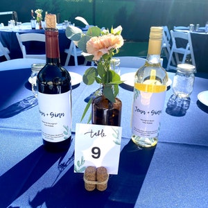 May include: A blue tablecloth with two bottles of wine labeled "Jenn + Gian" and a table number card with the number 9. The bottles are surrounded by greenery and a vase with a pink flower.