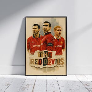 May include: Framed art print featuring three soccer players in red jerseys. The players are depicted in a painterly style, with the text "THE RED DEVILS" at the bottom. The artwork is set against a neutral background.