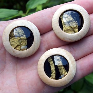 May include: Three round wooden buttons with a black center and gold leaf accents are displayed in a hand. The buttons have a light wood border. The background is blurred green foliage.