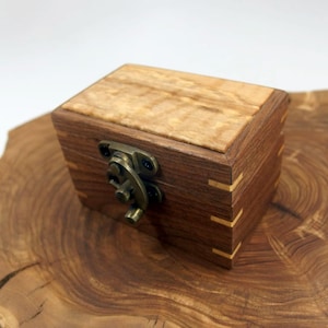 May include: A small wooden box with a hinged lid and a brass latch. The box is made of light and dark wood, and the lid has a natural wood grain pattern.