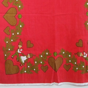 May include: Red tablecloth with a white border featuring a repeating pattern of brown hearts, green holly sprigs, and a couple of figures in green and brown clothing.