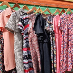 May include: A rack of vintage dresses in various colors and patterns, including pink, black, white, red, and floral prints. The dresses are hanging on hangers and are ready to be worn.