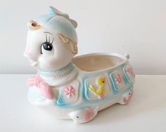 May include: A ceramic planter shaped like a turtle with a blue hat, pink bow, and yellow bird design. The planter is white with blue, pink, and yellow accents.