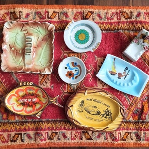 May include: A collection of six colorful ceramic ashtrays. The ashtrays feature various designs, including a sunflower, a geometric pattern, a lobster, and a depiction of the "Great Meteor Crater" in Arizona.