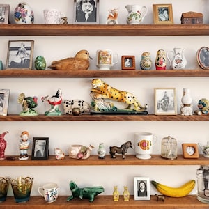 May include: A shelf with a variety of decorative items, including figurines, mugs, and framed pictures. There are several animal figurines, including a leopard, a duck, and a bird. There are also several mugs, including a white mug with a floral design and a blue mug with a white design. The shelf is made of wood and has a light brown finish.