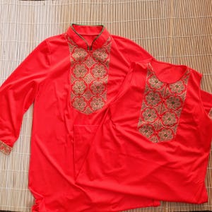 May include: Two-piece red outfit with gold and green embroidered floral patterns. The long-sleeved top has a mandarin collar with green trim. The sleeveless top has a V-neck. Both pieces are displayed on a bamboo mat.