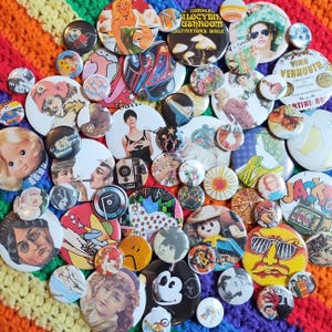 May include: A collection of colorful vintage button pins with various designs, including a mushroom, a car, a woman, a face, and a flower. The buttons are arranged in a random pattern on a rainbow crocheted surface.