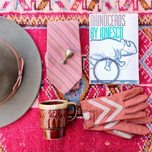 May include: An assortment of vintage items on a pink rug. Includes a gray hat, a striped tie, a small brass cup, a brown mug, a book titled "Rhinoceros by Ionesco", a bracelet, and pink gloves with white chevron details.