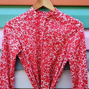 May include: A red and white floral print dress with long sleeves, a mandarin collar, and a button-down front. The dress is hanging on a wooden hanger. The fabric is covered in small red flowers.