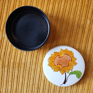 May include: A black round trinket box with a white lid featuring a yellow sunflower with a brown center and green leaves.