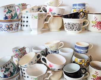 May include: A collection of ceramic mugs and cups featuring various designs, including floral patterns, geometric shapes, and a Japan-themed graphic. The assortment includes white, black, blue, and yellow pieces with diverse handle styles.