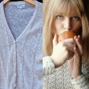 May include: A light beige, open-weave cardigan with a V-neck and button closure. The sweater features a cable knit design along the front edges and sleeves. The label reads "Atelier Fane".