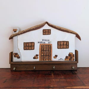 May include: A white ceramic wall-mounted key holder shaped like a house with a thatched roof, a chimney, and brown accents. The front features the text "VILLA MARIA" above a central door, with four metal hooks attached to a wooden base below.